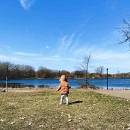 A young child in a brown jacket with the hood over their head walks near the water alone.